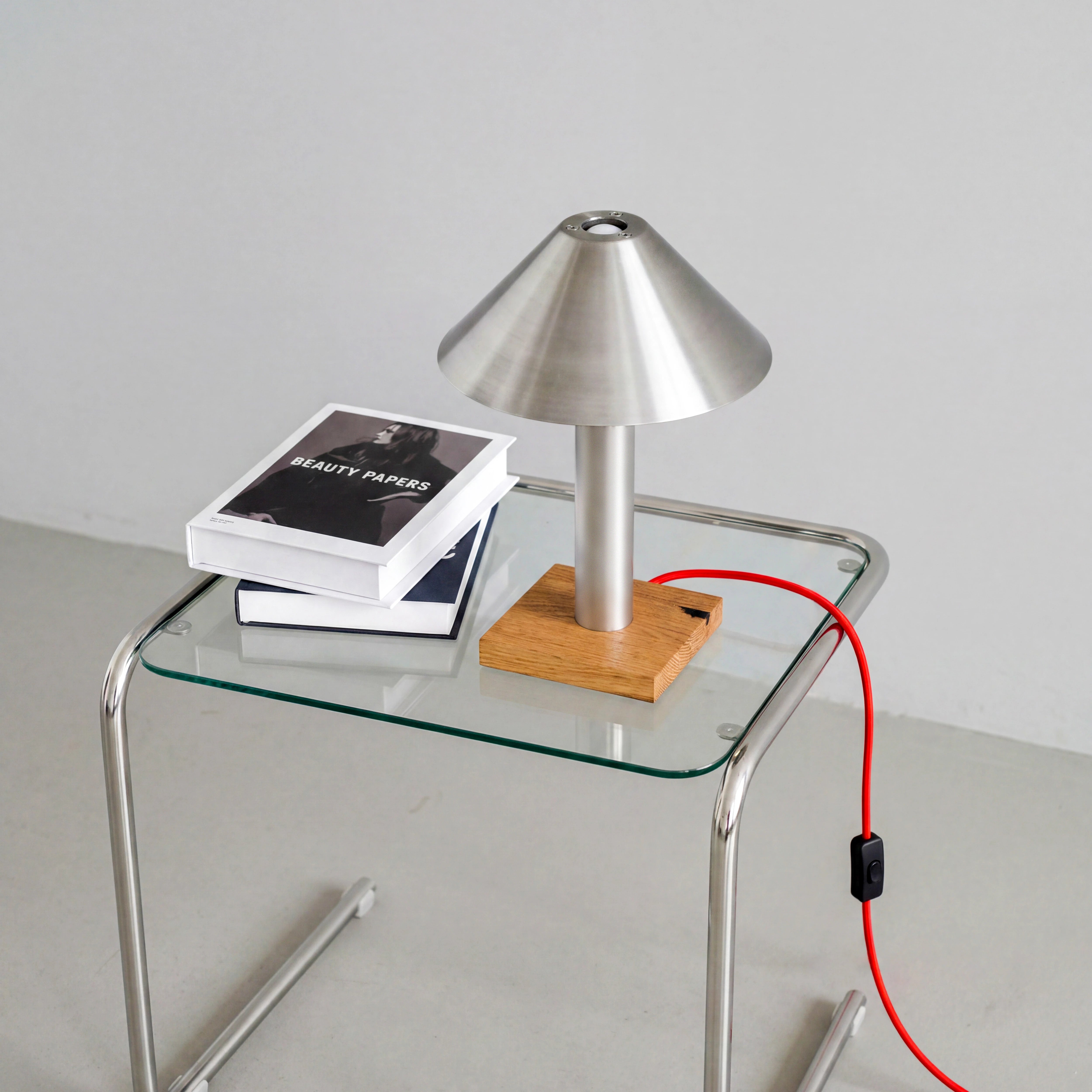 Tischleuchte PFEIL aus Edelstahl und Eiche auf einem Beistelltisch aus Glas und Chrom, mit rotem Textilkabel und Kippschalter.
