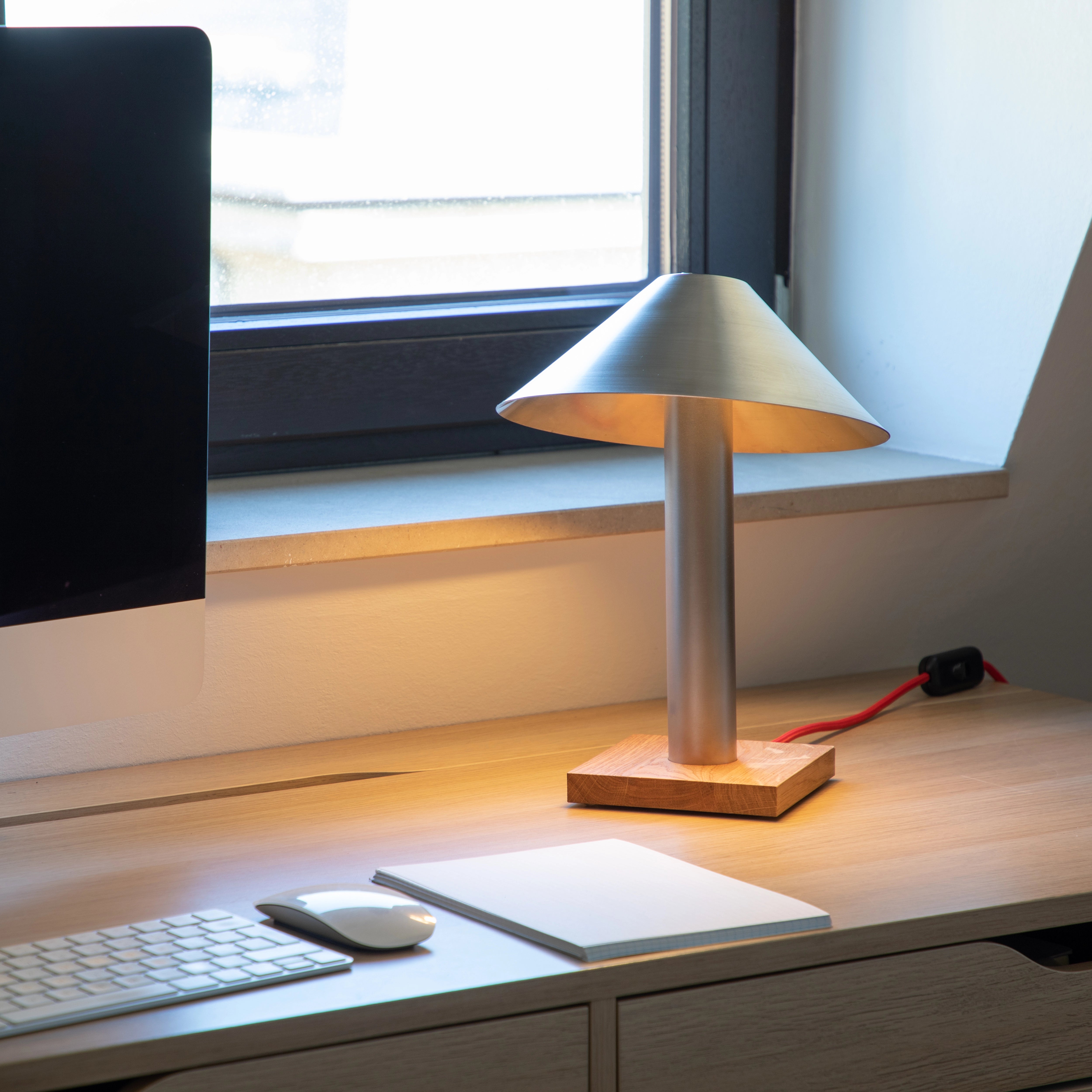 Leuchtende Schreibtischlampe PFEIL aus Edelstahl mit Eichensockel am Fenster neben Monitor und Tastatur
